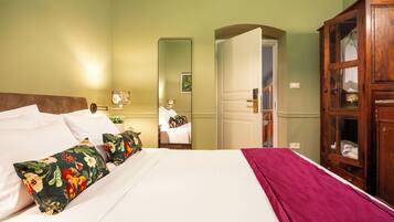 A neatly made bed with floral pillows, a wooden wardrobe, and a mirror reflecting a room with a door and a plant.