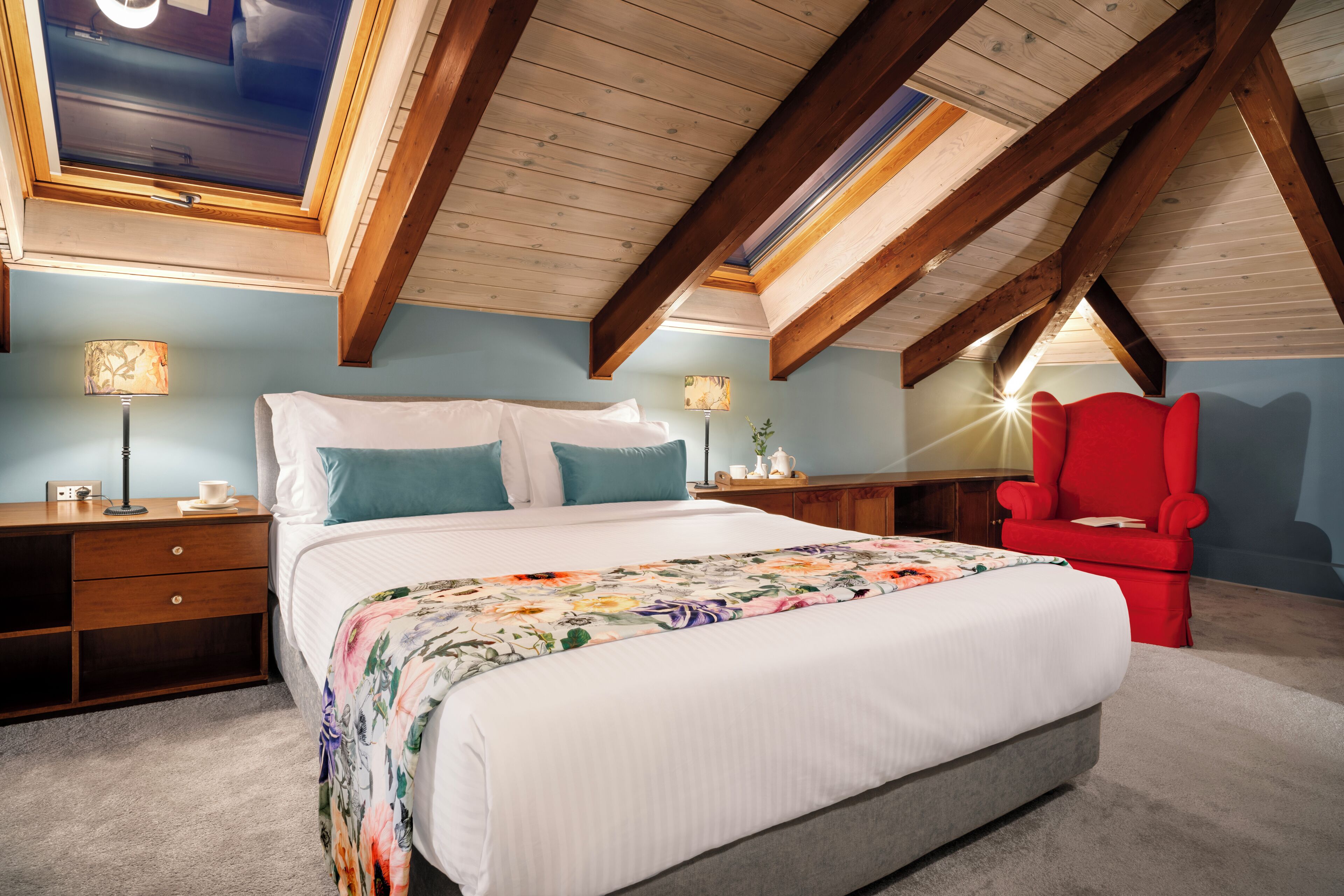 Een slaapkamer met een groot bed, een rode stoel, houten balken, een dakraam en een nachtkastje met een lamp.