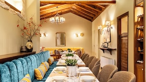 Un comedor con una larga mesa dispuesta para una comida, un banco tapizado en azul con almohadones amarillos, y una araña colgante.