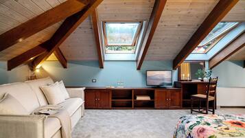 Una habitación con techo inclinado, vigas de madera, un sofá, un escritorio con una silla, un televisor y un tragaluz.