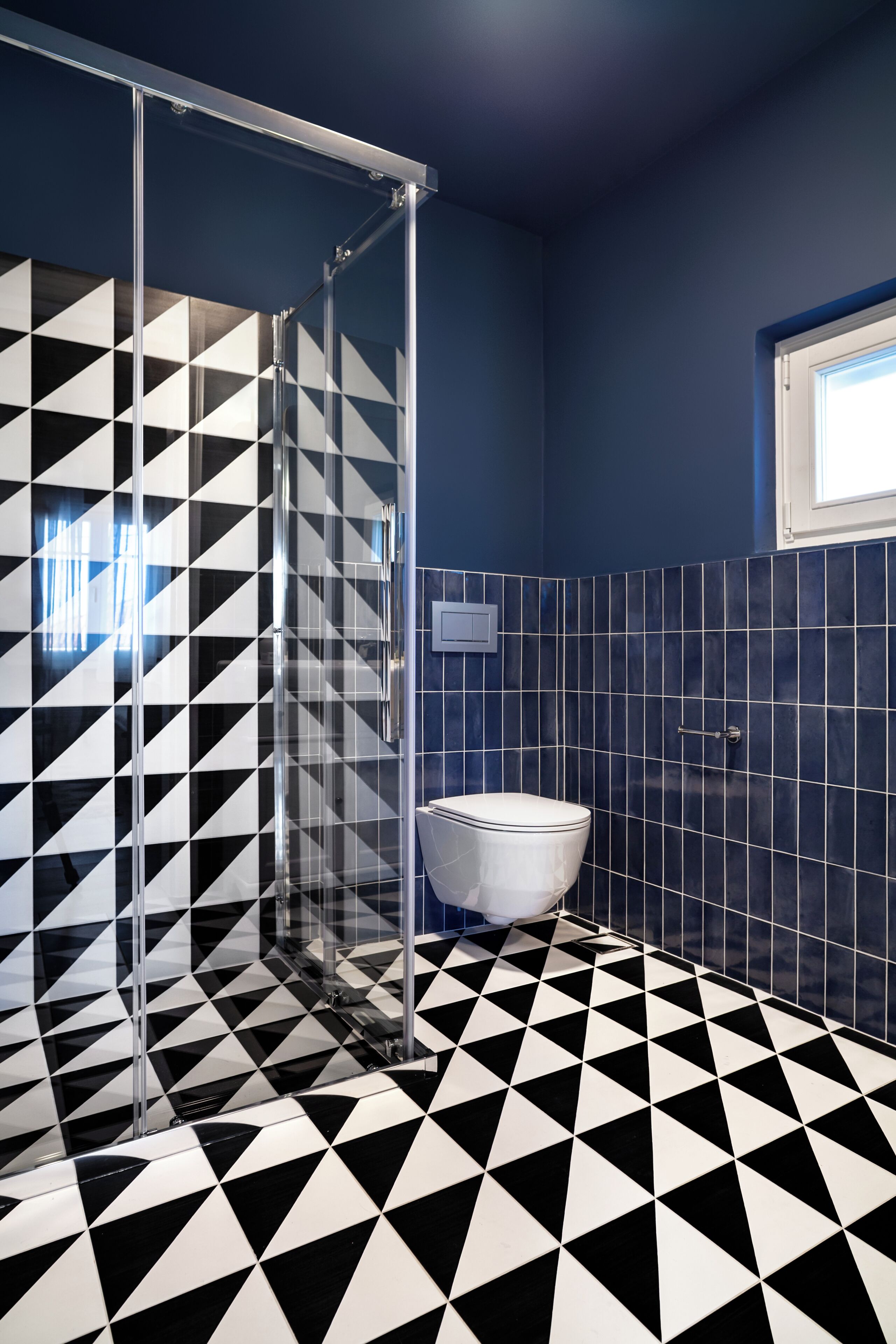 Un bagno con pavimento a scacchi bianchi e neri, cabina doccia in vetro e un water su una parete rivestita di piastrelle blu.