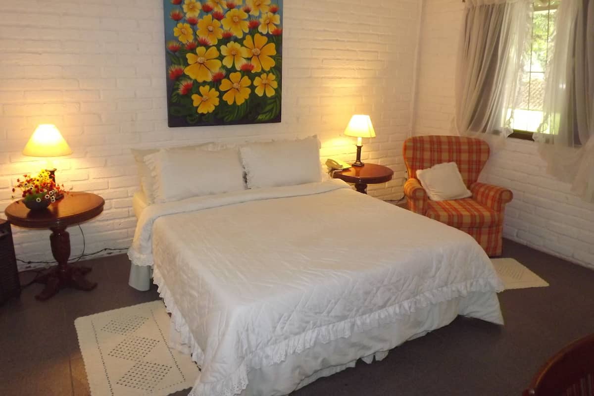 Ein Schlafzimmer mit einem großen Bett, zwei Nachttischen mit Lampen, einem karierten Sessel, einem Blumenbild an der Wand und einem Fenster mit Vorhängen.