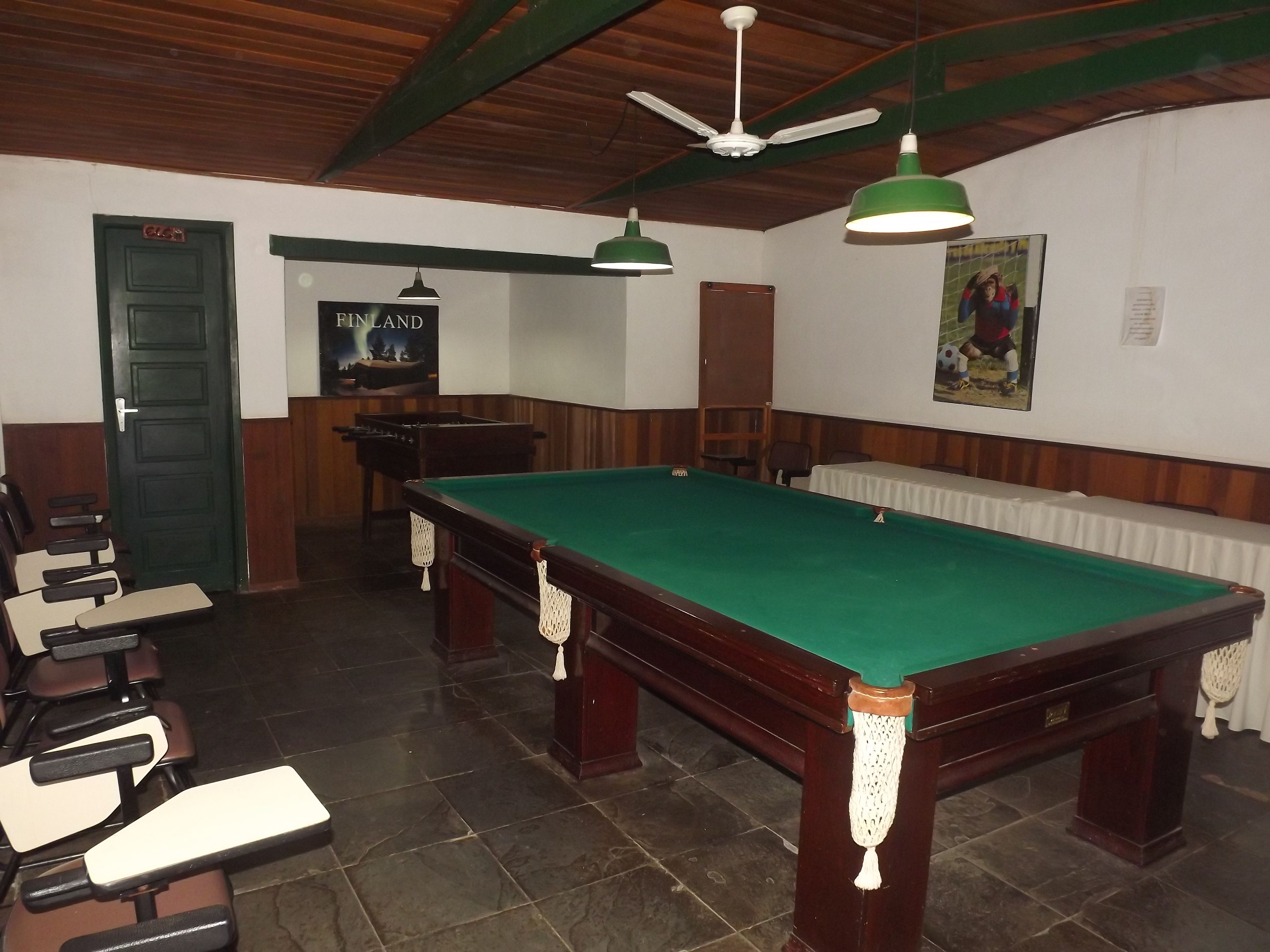 Uma sala de bilhar com mesa de sinuca, cadeiras, um cartaz e um ventilador de teto.