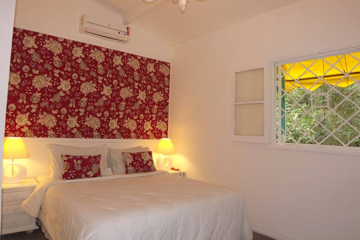 Ein Schlafzimmer mit einem blumengemusterten Kopfteil, einem Bett mit weißer Bettwäsche, zwei Nachttischlampen, einem Fenster mit gelbem Vorhang und einer Klimaanlage an der Wand.