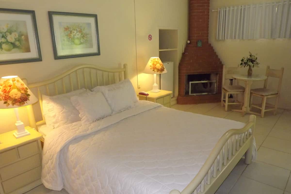 Ein Schlafzimmer mit einem großen Bett, zwei Nachttischlampen, einem Kamin, einem kleinen Tisch mit Stühlen und zwei gerahmten floralen Bildern an der Wand.