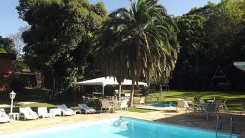 Relax by the serene pool surrounded by lush greenery and palm trees, with comfortable seating and a shaded area.