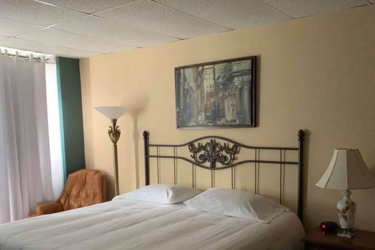 Una cama bien hecha con cabecera, un cuadro en la pared, una lámpara en la mesita de noche y una silla en un rincón.