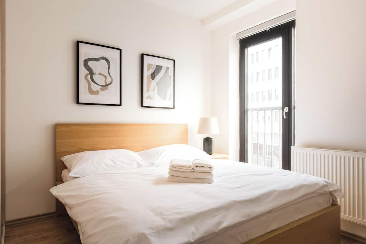 A neatly made bed with white linens and a stack of folded towels, a bedside lamp, and two abstract art pieces on the wall.
