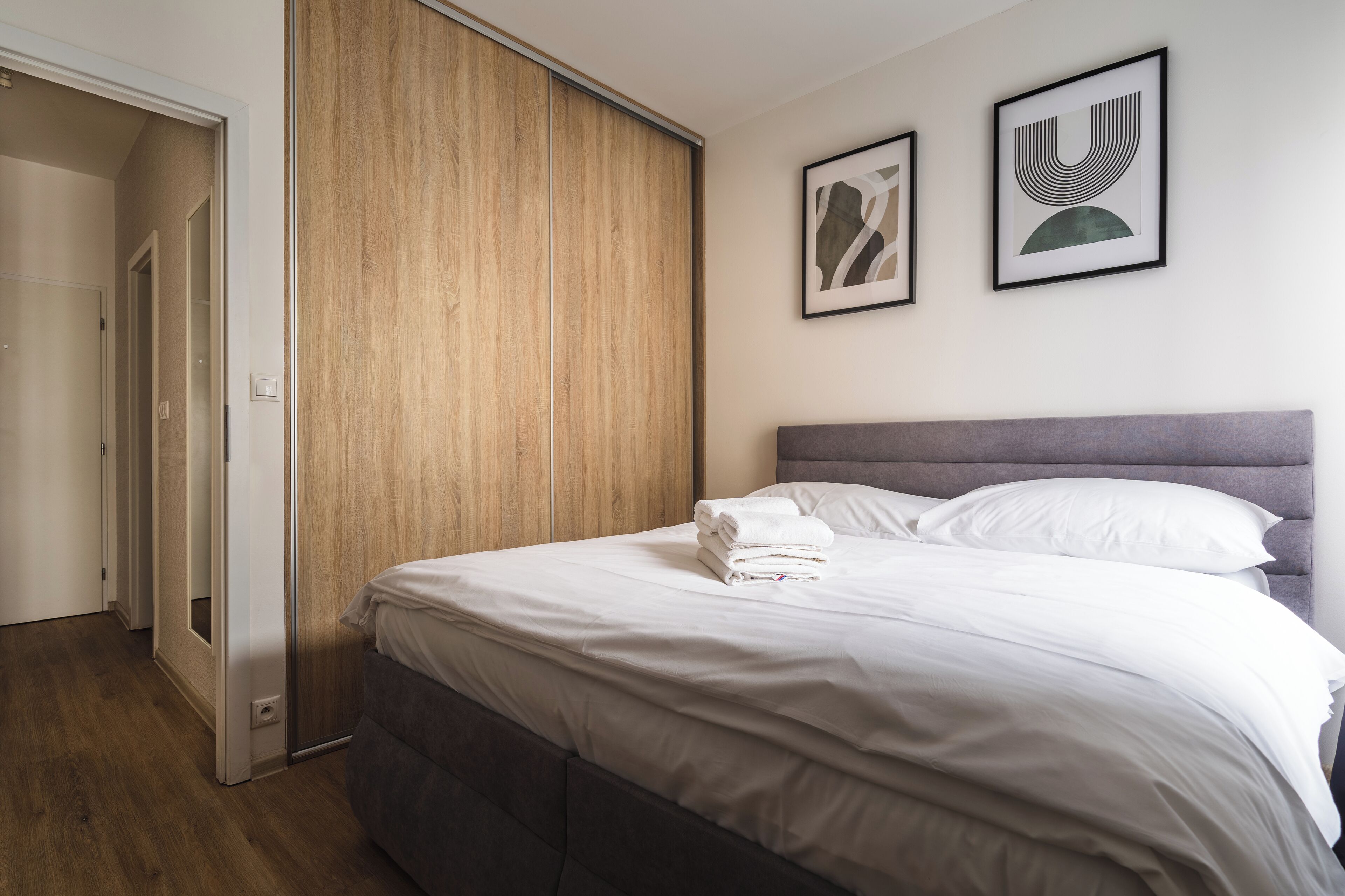 A bedroom with a bed, wooden wardrobe, and two framed artworks on the wall.