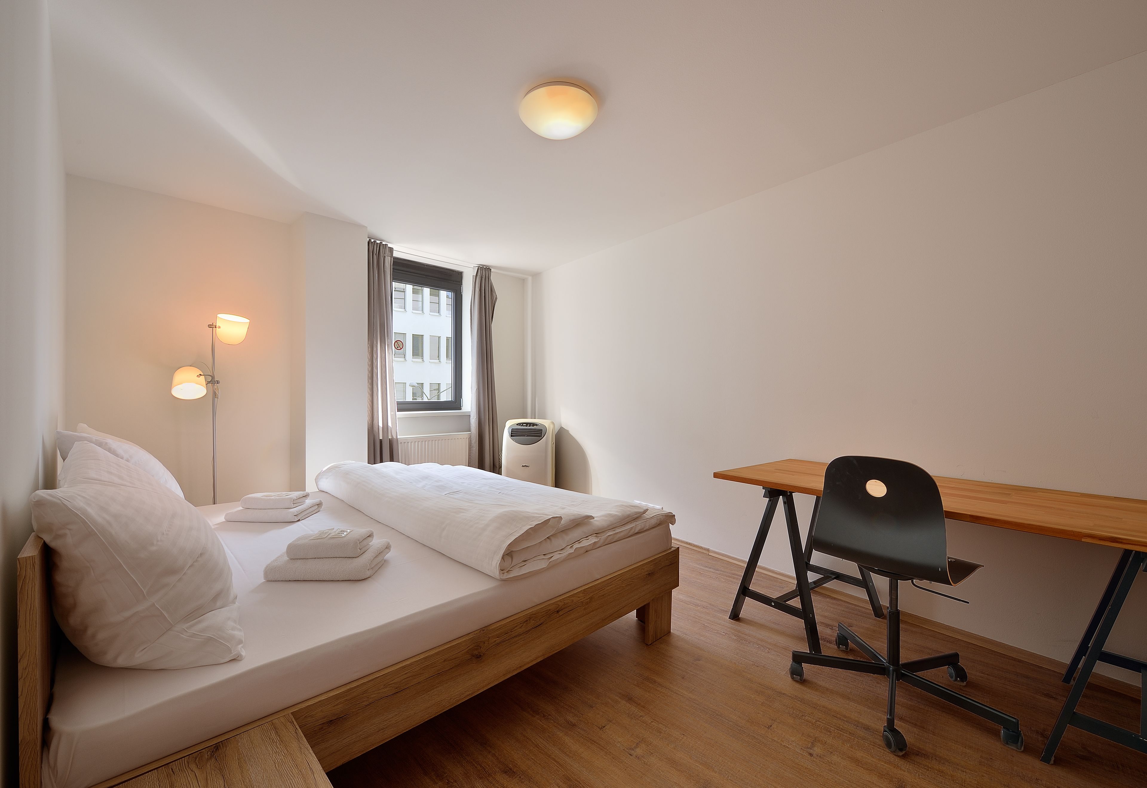 A bedroom with a wooden bed, a desk, a chair, a window with curtains, and a wall-mounted light fixture.