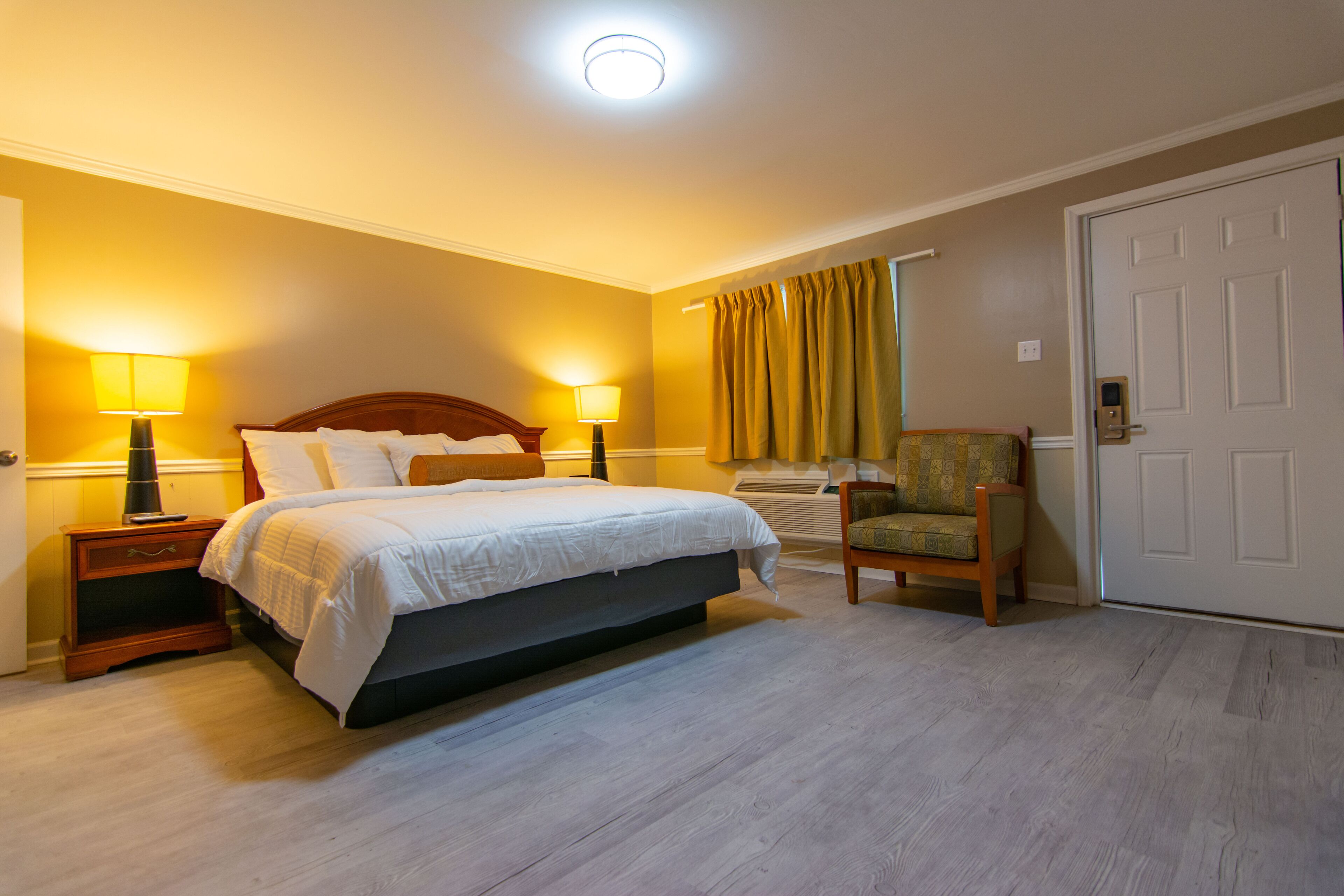 Une chambre d’hôtel avec un lit, deux lampes de chevet, une chaise, une porte et une fenêtre avec des rideaux.