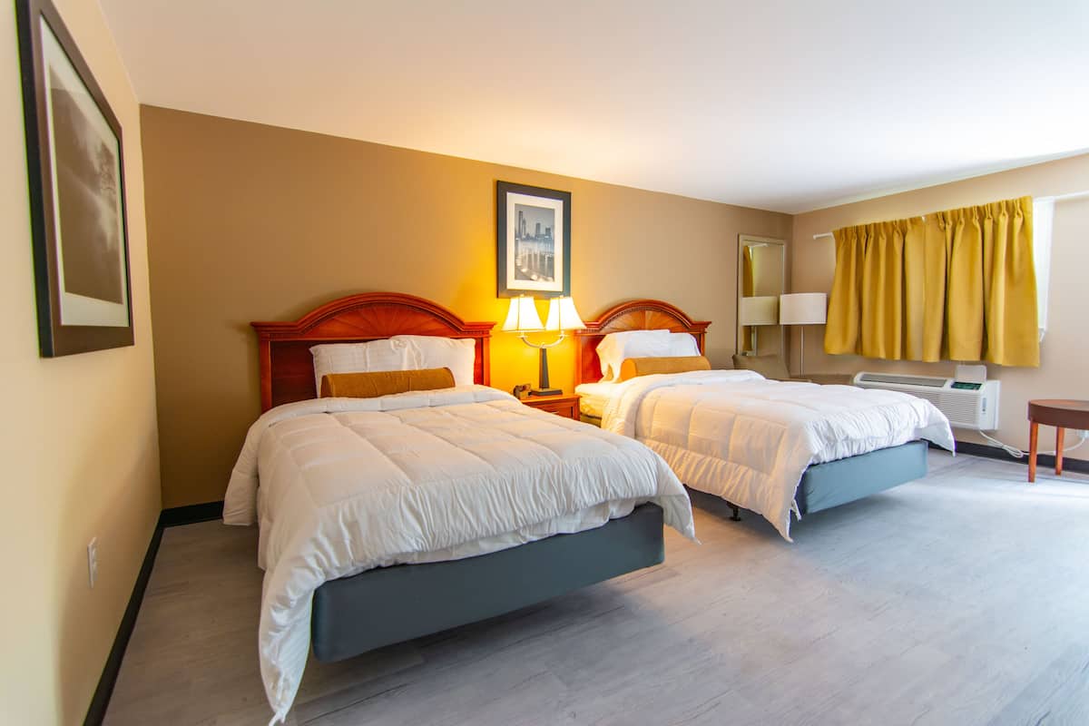 Quarto de hotel com duas camas, mesa de cabeceira com luminárias, espelho, janela com cortinas amarelas e um quadro na parede.