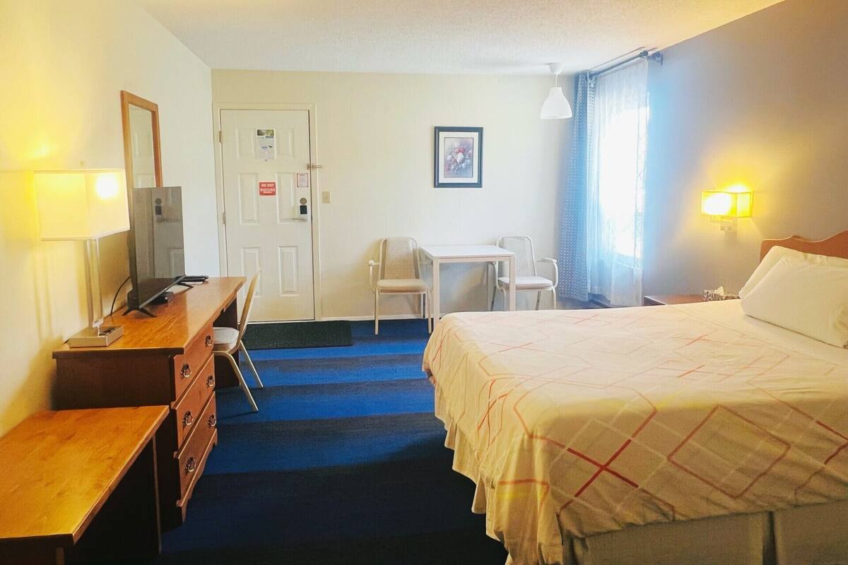 A hotel room with a bed, desk, chair, dresser, and a door with a sign.