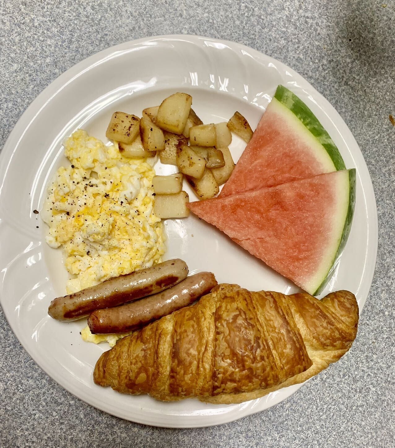Un plato con huevos revueltos, patatas asadas, salchichas, una rebanada de sandía y un croissant.