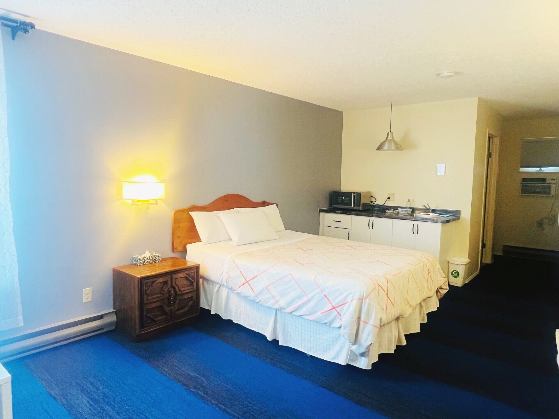 A hotel room with a bed, bedside table, lamp, kitchenette, and a blue carpet.