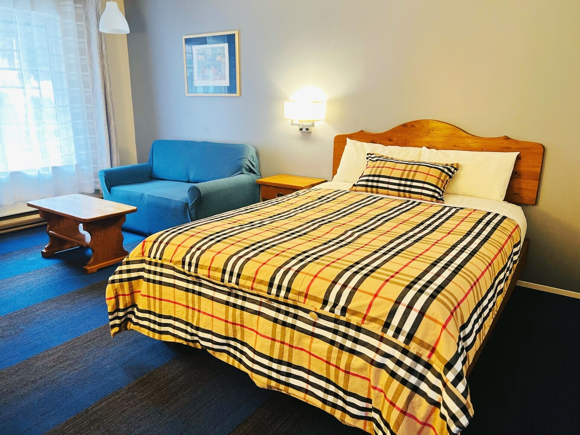 Een hotelkamer met een bed, een blauwe fauteuil, een klein houten tafeltje, een nachtkastje en een wandlamp.