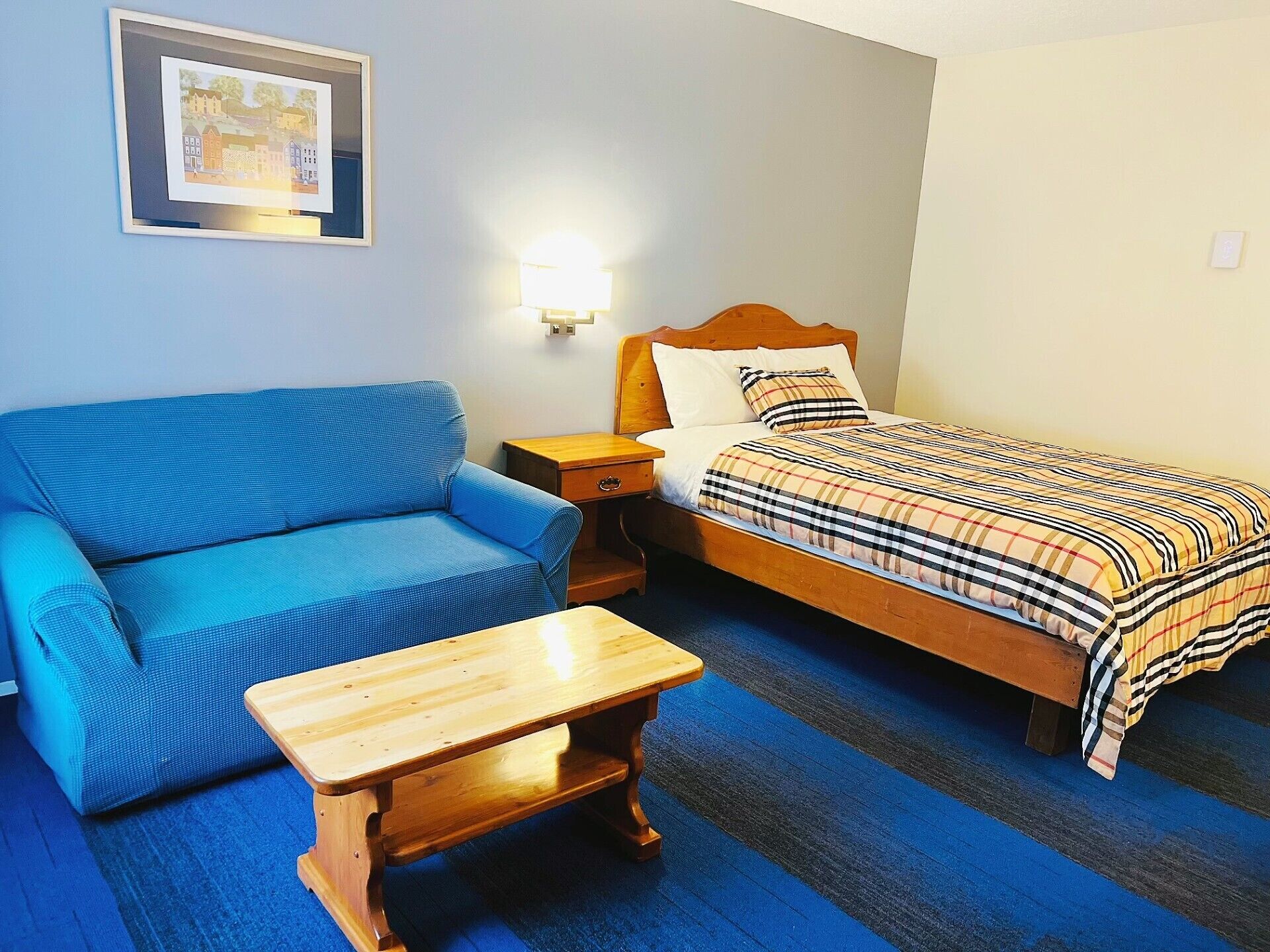Una habitación con un sofá azul, una cama de madera con una manta escocesa, una mesita de madera y una lámpara fijada a la pared.