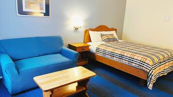 Una habitación con un sofá azul, una cama de madera con una manta escocesa, una mesita de madera y una lámpara fijada a la pared.