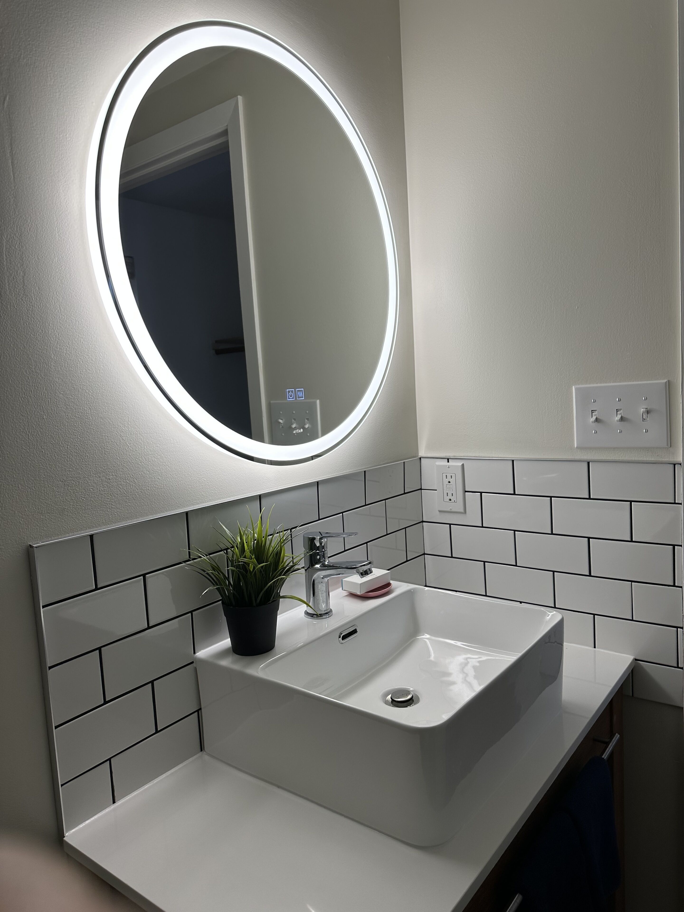 Un baño con un lavamanos blanco, un espejo redondo con luz y una planta en maceta.