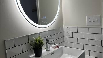 Un baño con un lavamanos blanco, un espejo redondo con luz y una planta en maceta.