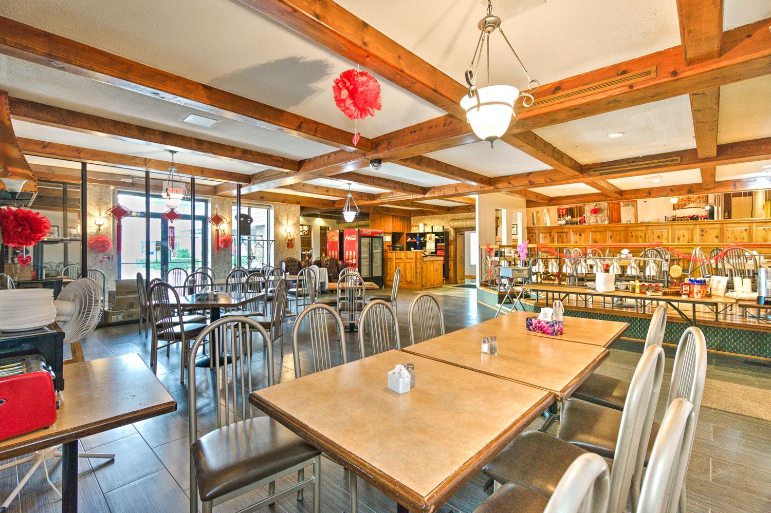 A spacious restaurant with wooden beams, multiple tables and chairs, and a Coca-Cola refrigerator.