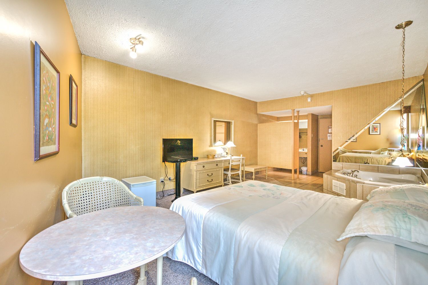 Una habitación de hotel con cama, escritorio con silla, una mesita, televisor y baño con espejo y lavamanos.