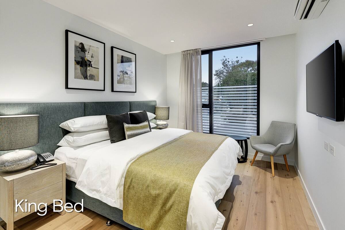 Una cama tamaño king con un plaid verde, una lámpara de noche y un teléfono sobre una mesita de madera. Hay una silla gris y un televisor montado en la pared.