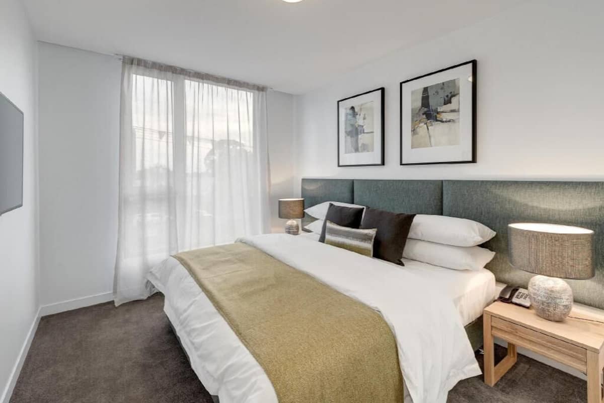 Habitación de hotel con cama, lámparas de noche, mesita de noche, televisor montado en la pared y dos cuadros enmarcados.