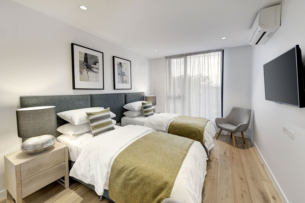 Een moderne hotelkamer met twee bedden, een nachtkastje, een stoel, een flatscreen-tv en kunst aan de muur.