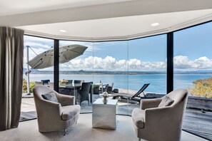 A modern living room with a large glass wall offering a scenic view of the sea and clouds. - Serenity on Wakeman (Taupo)