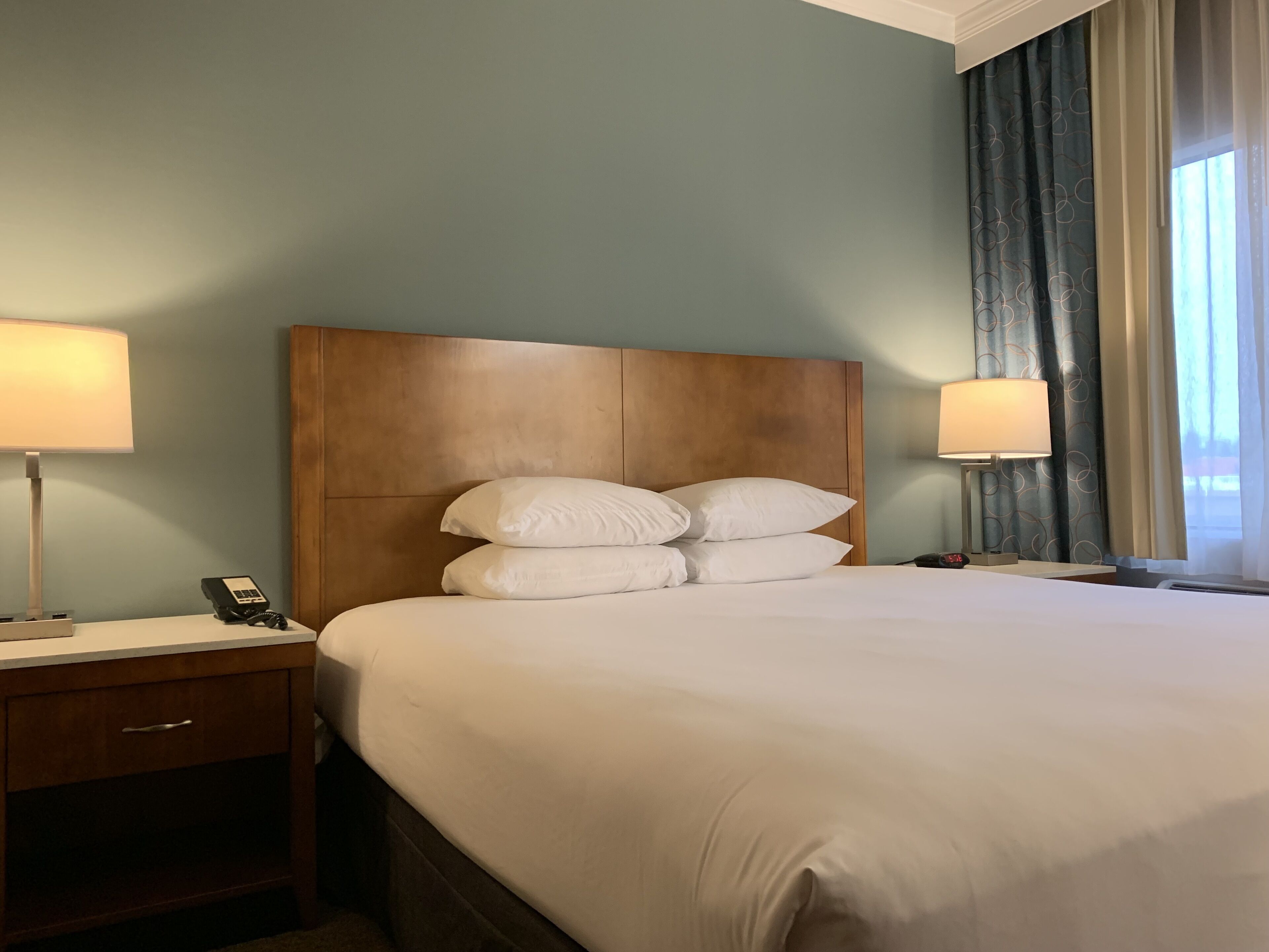 Une chambre d’hôtel avec une tête de lit en bois, un grand lit avec du linge de lit blanc, deux tables de chevet avec des lampes, et une vue sur l’extérieur par une fenêtre avec des rideaux.