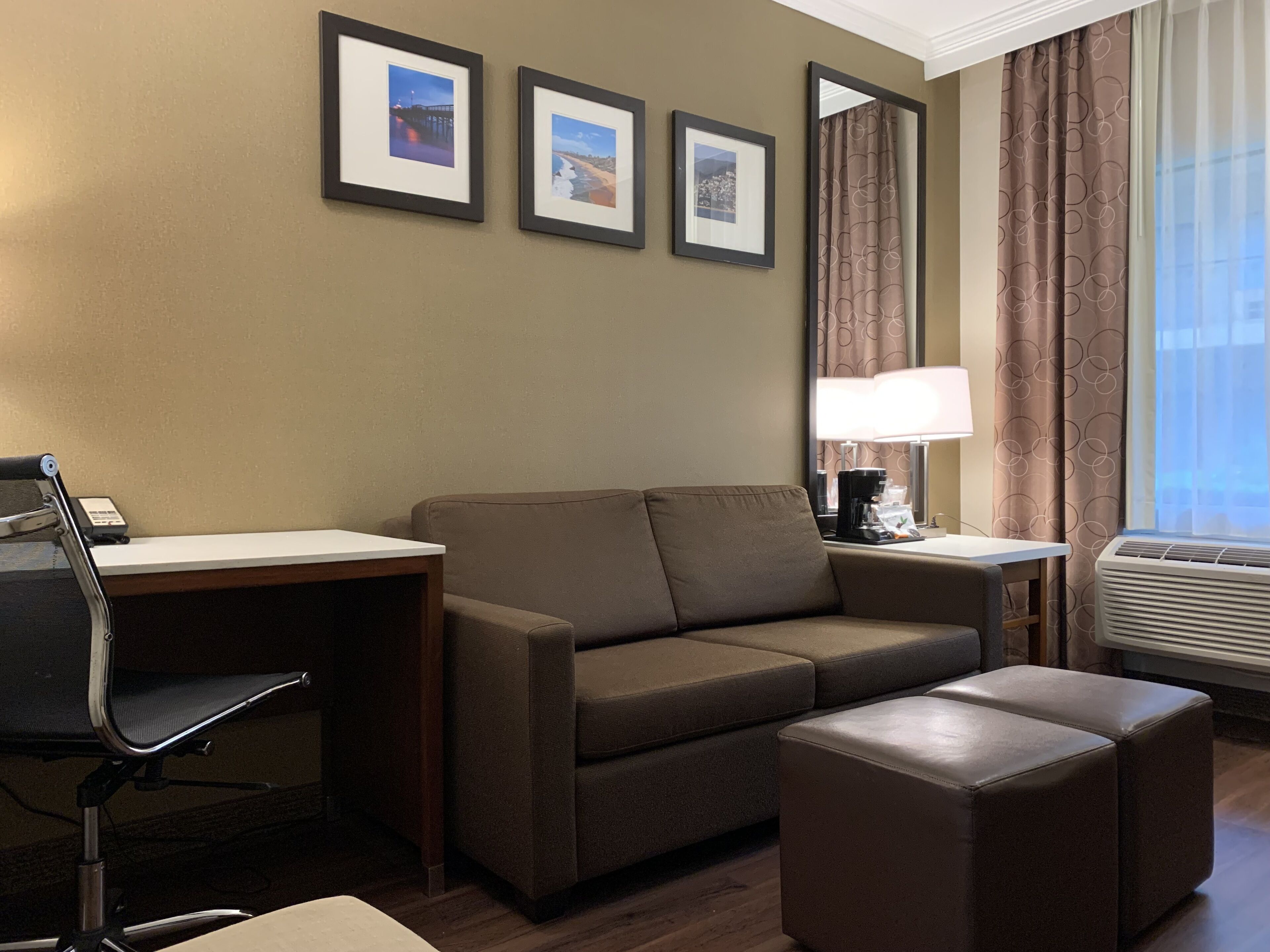 A hotel room with a sofa, a desk, a chair, an ottoman, a lamp, and framed pictures on the wall.