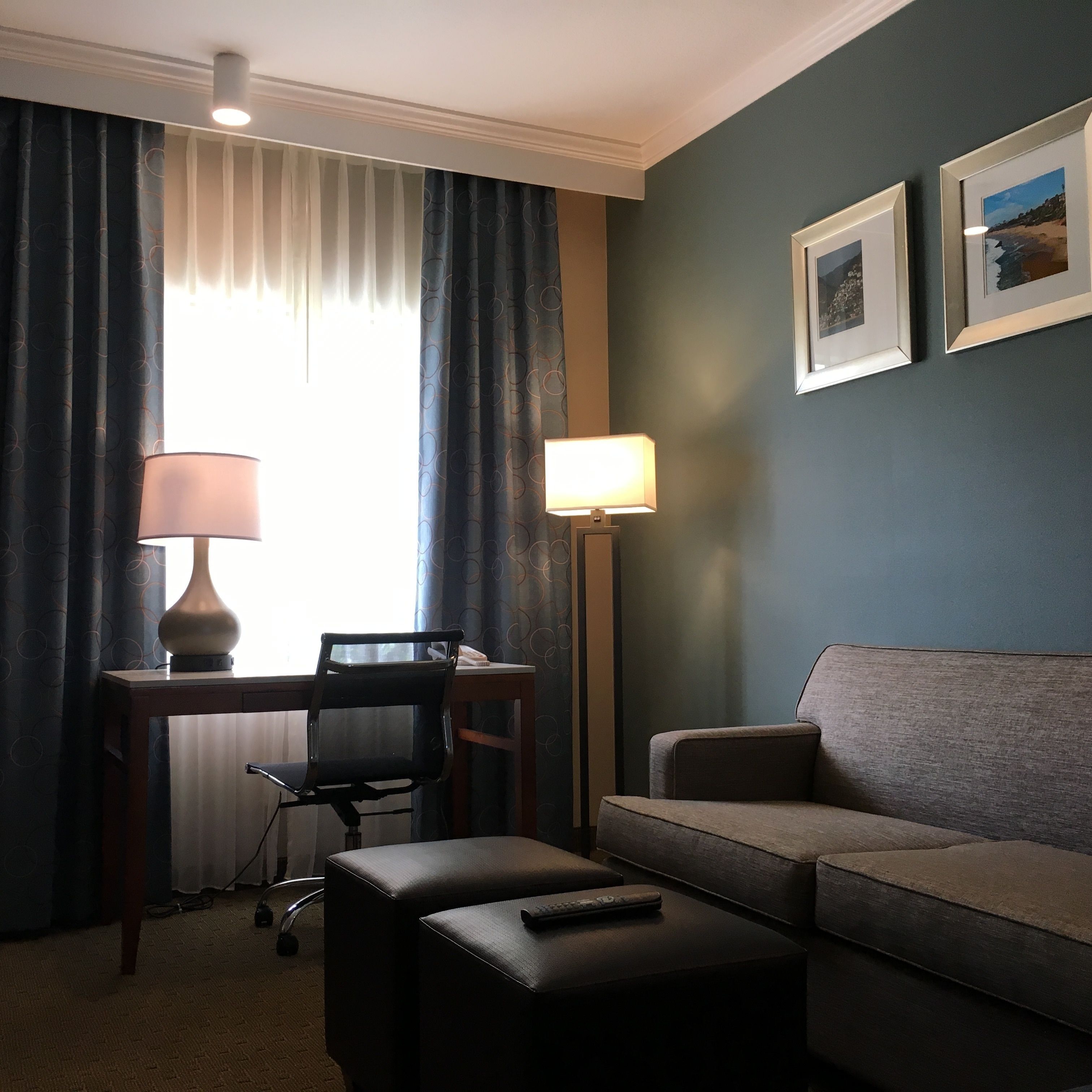 A hotel room with a desk, two chairs, a sofa, a lamp, and a remote control on a table.