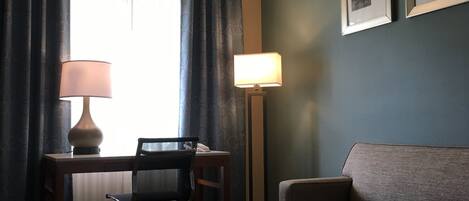 A hotel room with a desk, two chairs, a sofa, a lamp, and a remote control on a table.