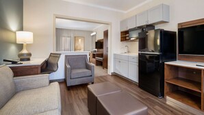 A modern hotel room with a sofa, armchair, ottoman, kitchenette, and a television.