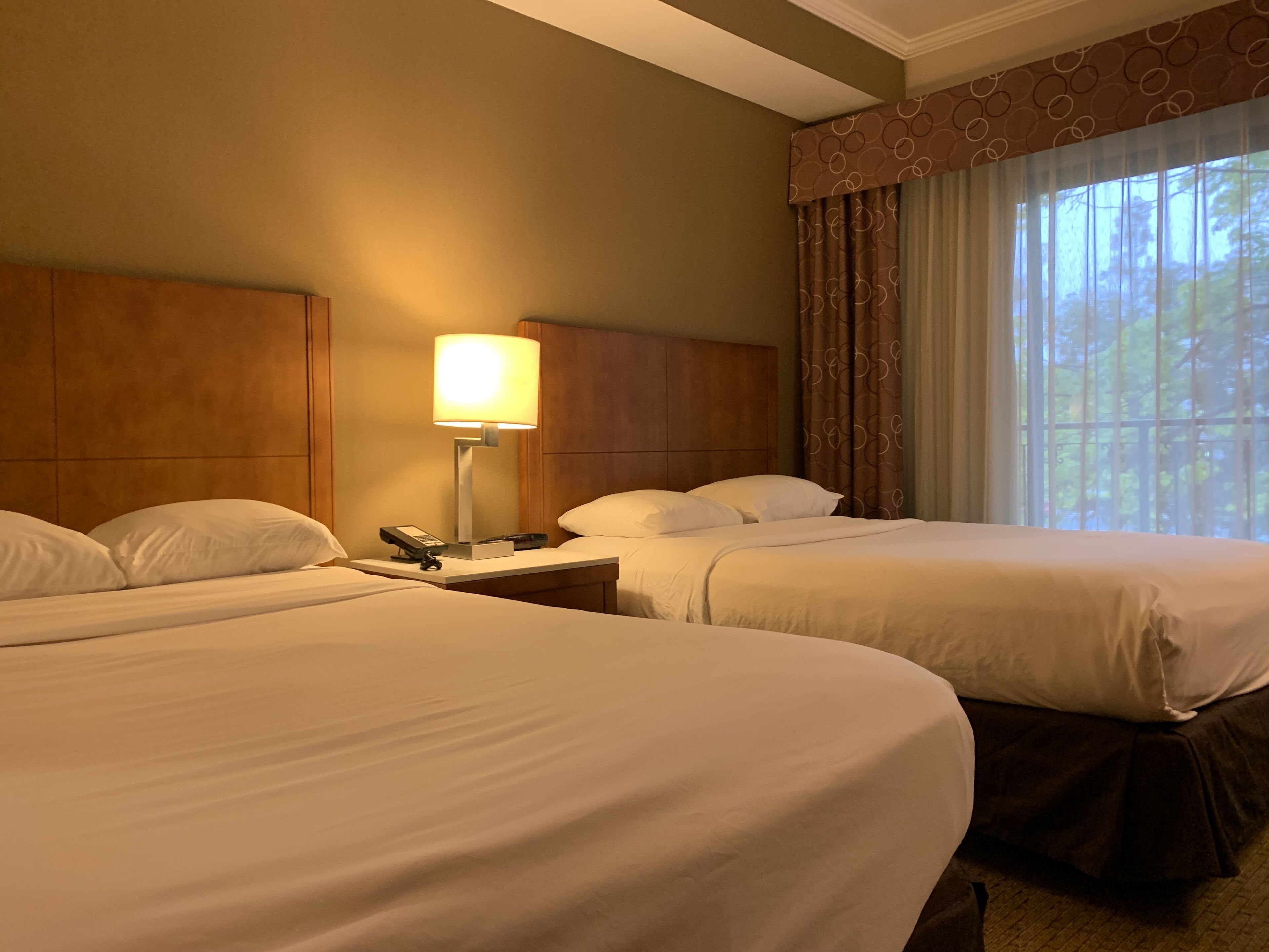 A hotel room with two beds, a bedside table with a lamp, a phone, and a window with curtains.