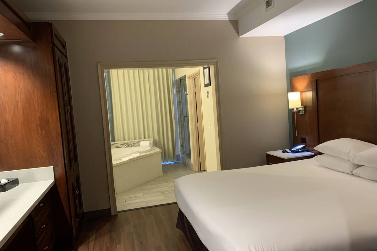 A hotel room with a bed, bedside table, lamp, and a bathroom with a shower and toilet.