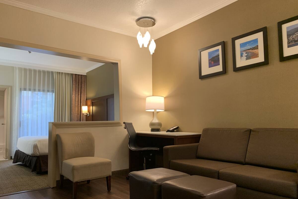 A hotel room with a bed, a desk, a chair, a sofa, and a small ottoman. There is a lamp on the desk and a mirror on the wall.