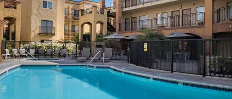 A swimming pool with a metal ladder, surrounded by a fence, and adjacent to a multi-story building with balconies and outdoor seating.