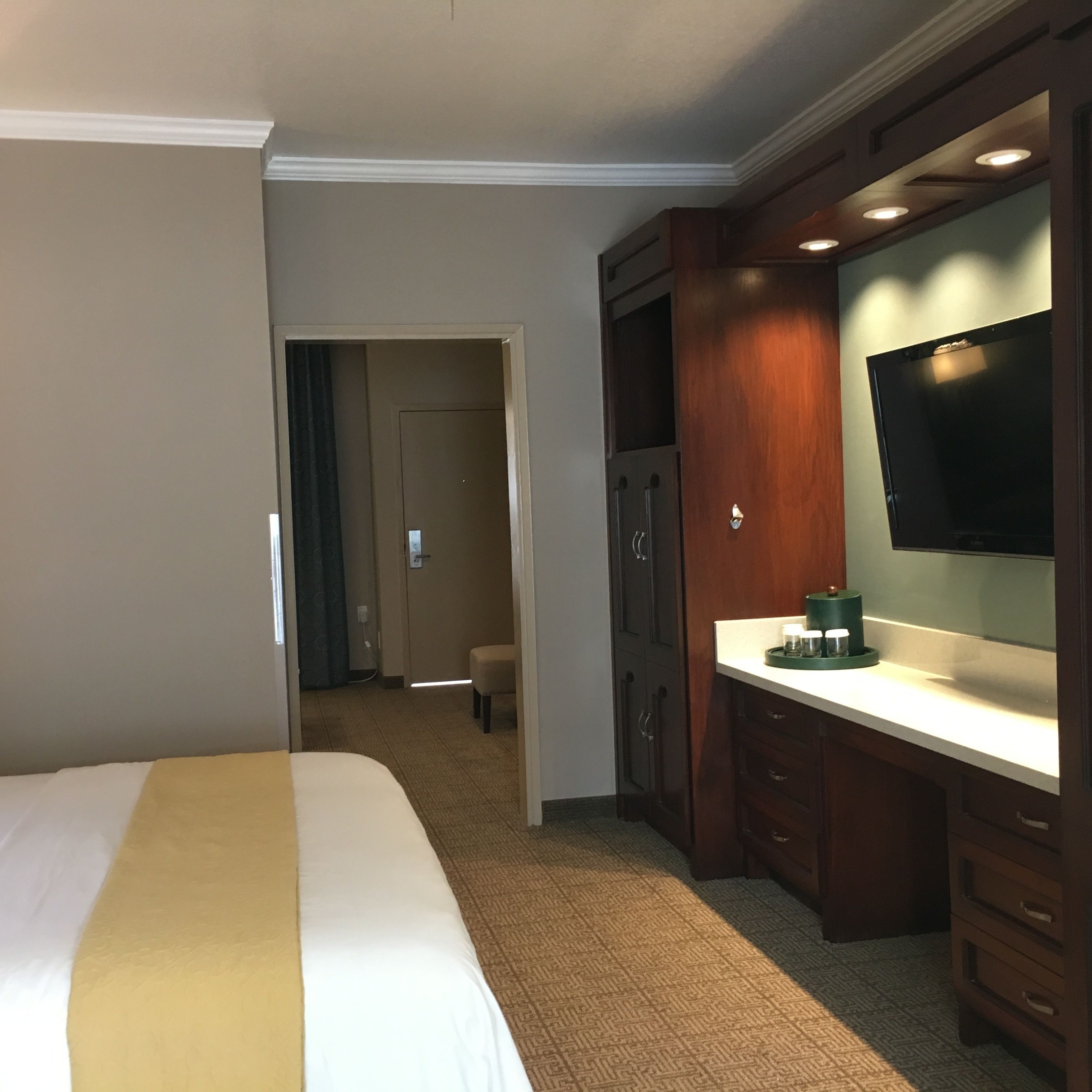 A hotel room with a bed, a desk with a flat-screen TV, a minibar, and a door leading to a bathroom.