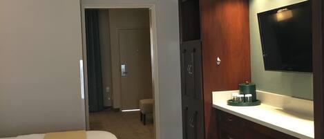 A hotel room with a bed, a desk with a flat-screen TV, a minibar, and a door leading to a bathroom.