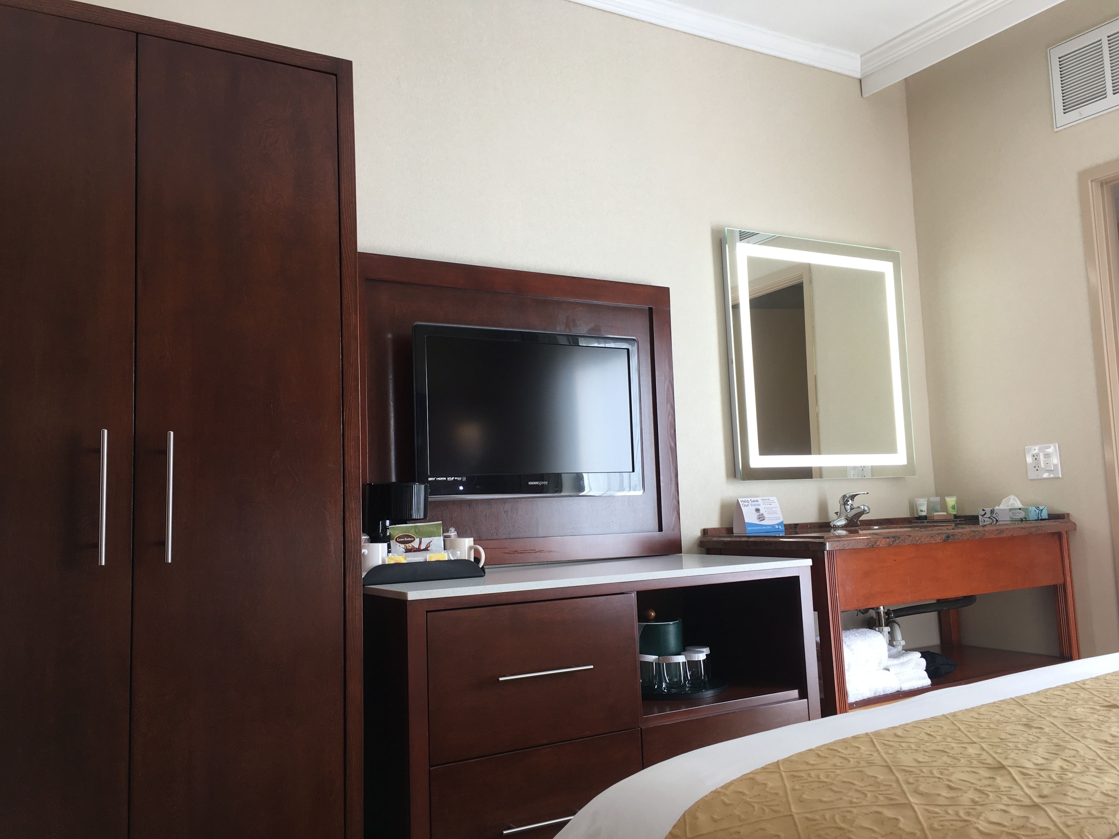A hotel room with a large wardrobe, a television, a mirror, a vanity with a sink, and a bed with a beige bedspread.