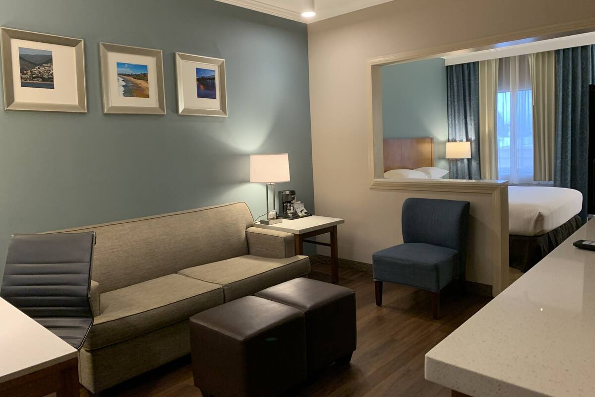 A hotel room with a sofa, a small table with a lamp, a chair, a bed, and a large mirror. There are three framed pictures on the wall.