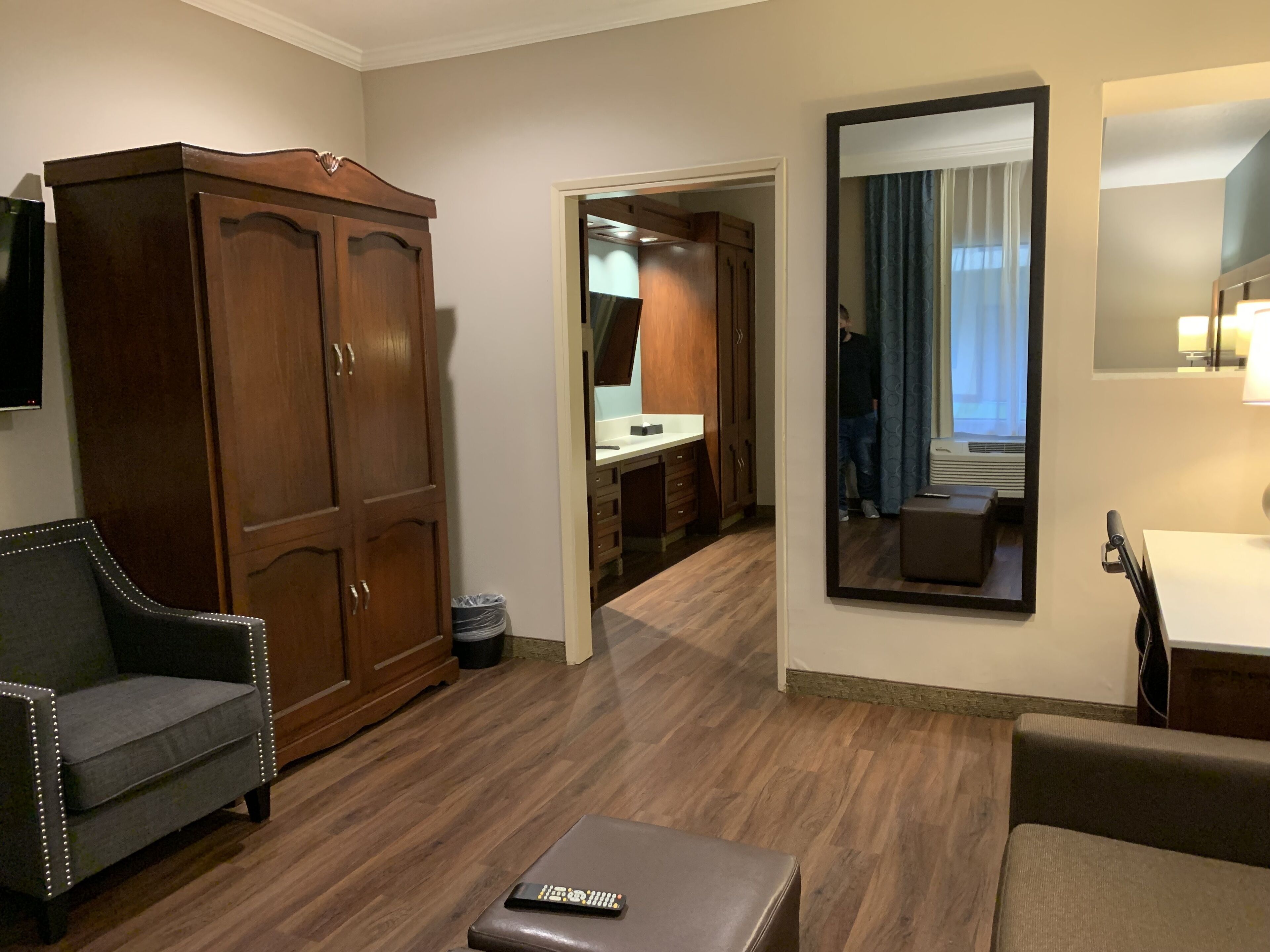 Habitación de hotel con un armario de madera, un sillón gris, un sofá marrón, una mesita pequeña y un espejo.
