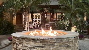 A stone fire pit with flames, surrounded by palm trees and a building with balconies in the background.