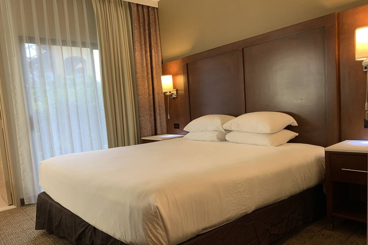 A hotel room with a large bed, a nightstand, a lamp, and a window with curtains.