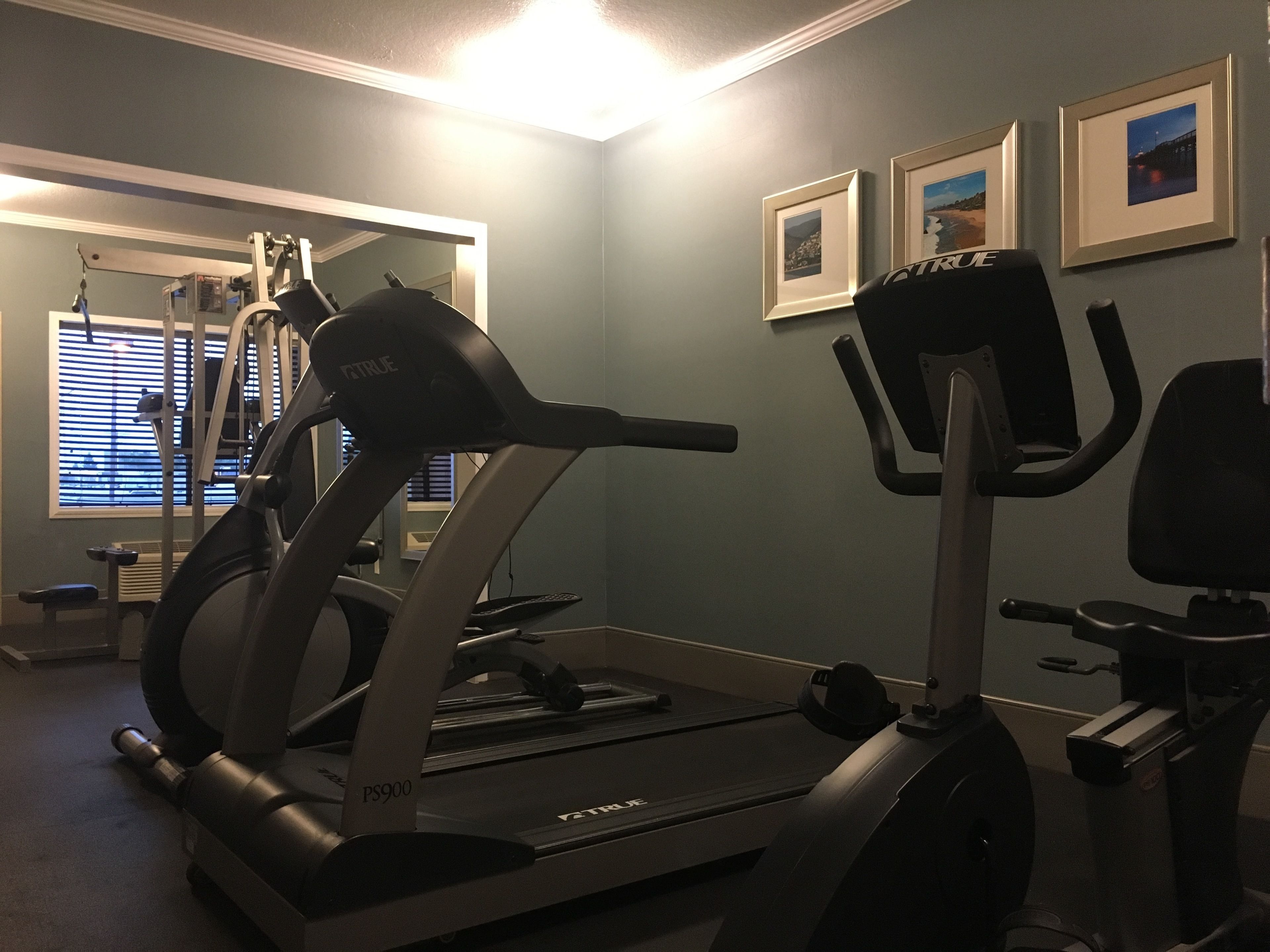 Ein Heimfitnessraum mit Laufband, Ergometer und Ellipsentrainer. An der Wand hängen Bilder und es gibt ein Fenster mit Jalousien.