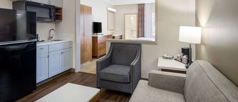 Una habitación de hotel compacta con kitchenette, un sillón gris, un sofá, una mesita y un espejo.