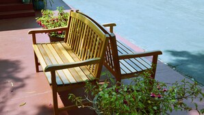 A wooden bench with a backrest, positioned on a paved surface next to a potted plant with green foliage and pink flowers. - Wittle Inn (Sunnyvale)