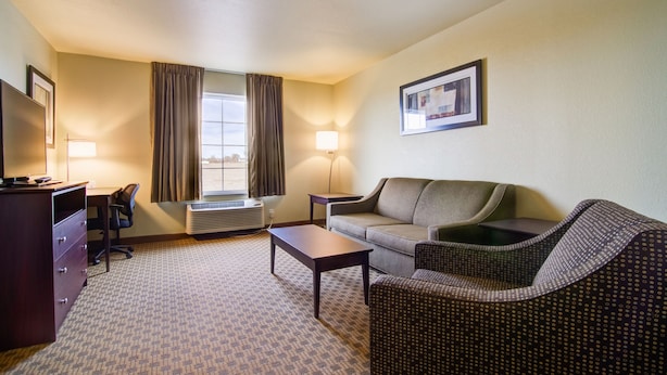 A hotel room with a desk, a sofa, a patterned rug, a window with curtains, and a framed picture on the wall. - Castle Rock Inn & Suites (Quinter)